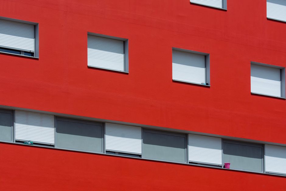 A sunlit urban building with red walls and closed shutters, showcasing architectural minimalism.